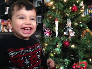 A boy smiles. The Christmas tree has ornaments including the Union Jack and Sherlock Holmes.