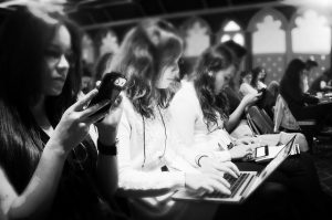 Female journalists seated at a news event, with one gathering information on her phone, another on her laptop, and a third on a tablet. Other women are seen in the background, but not in clear detail.