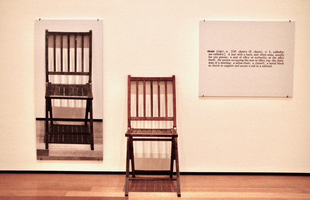 A wooden chair, with a picture of the same chair on the wall next to it, and a dictionary definition of the word "chair" on the wall on the other side of the chair.