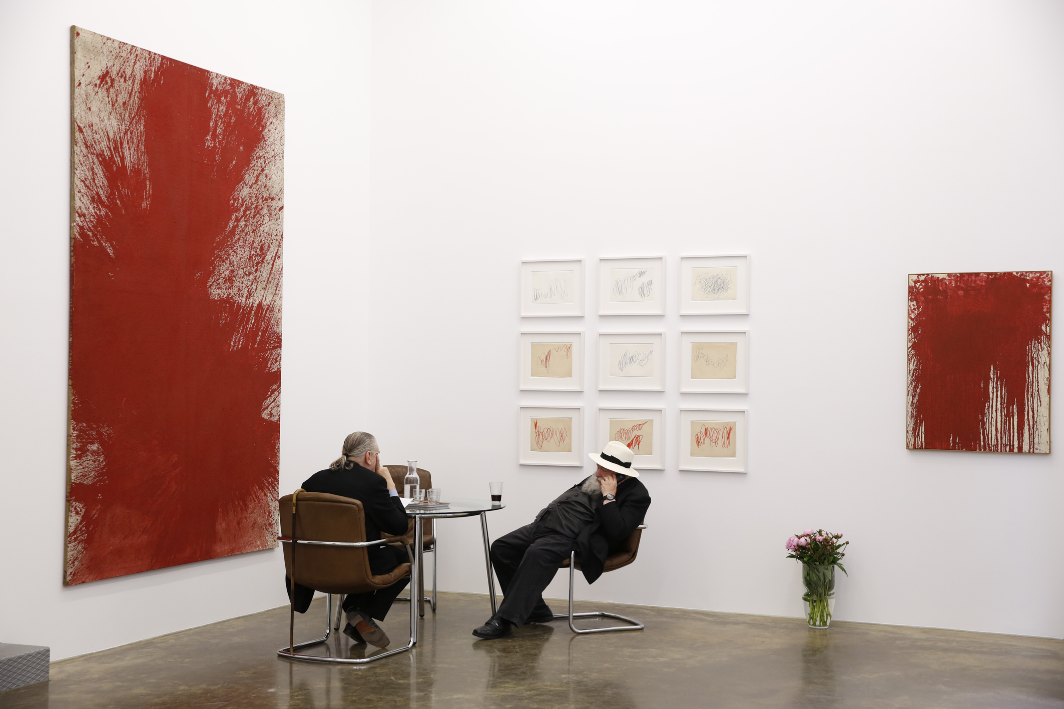 Two men sitting at a table with large paintings on the walls that have large red blotches of paint covering most of the canvas, and smaller drawings arranged in a 3 by 3 grid on part of the wall.