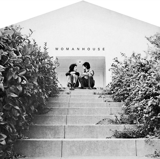 Grayscale image of two women sitting on the top flight of stairs leading to an entrance to a white-coloured house that has WOMANHOUSE letter-writing above.