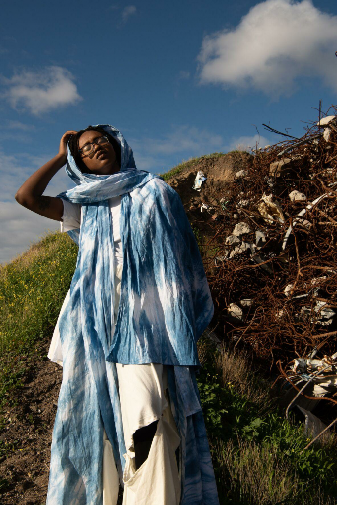 A woman with her eyes closed, face tilted towards the sky, with a green hillside on her right and a tangle of weeds and rocks on the same hillside on her left.