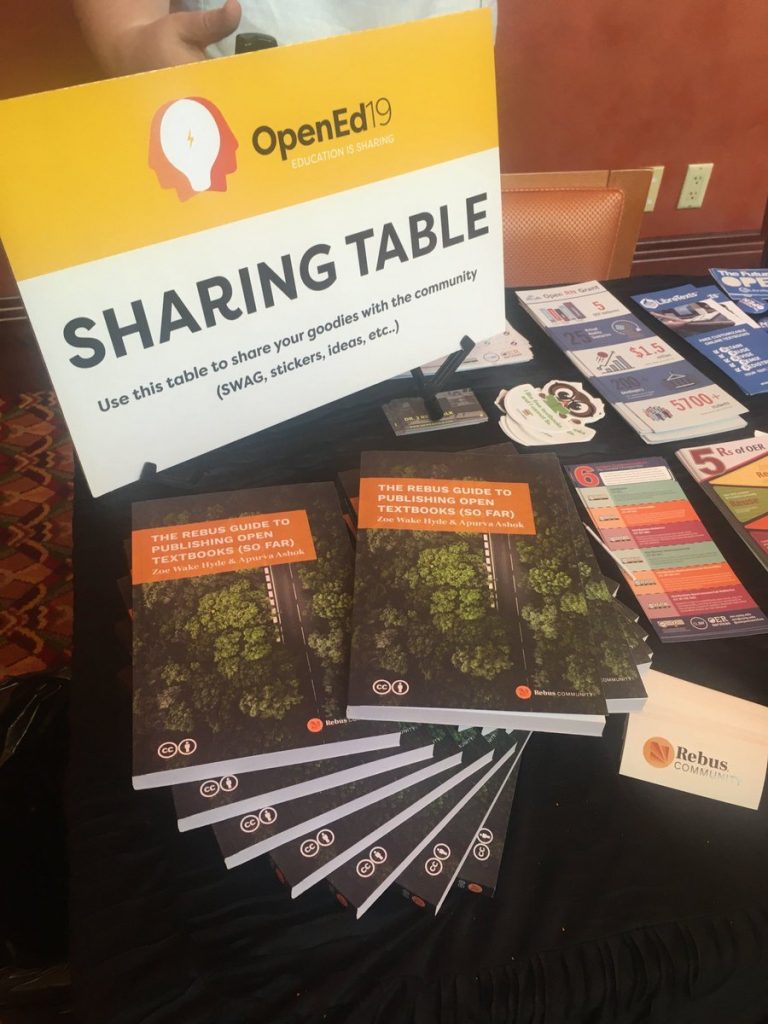 A sharing table at the 2019 Open Education conference with free items for attendees. The table includes print copies of “The Rebus Guide to Publishing Open Textbooks (So Far).”
